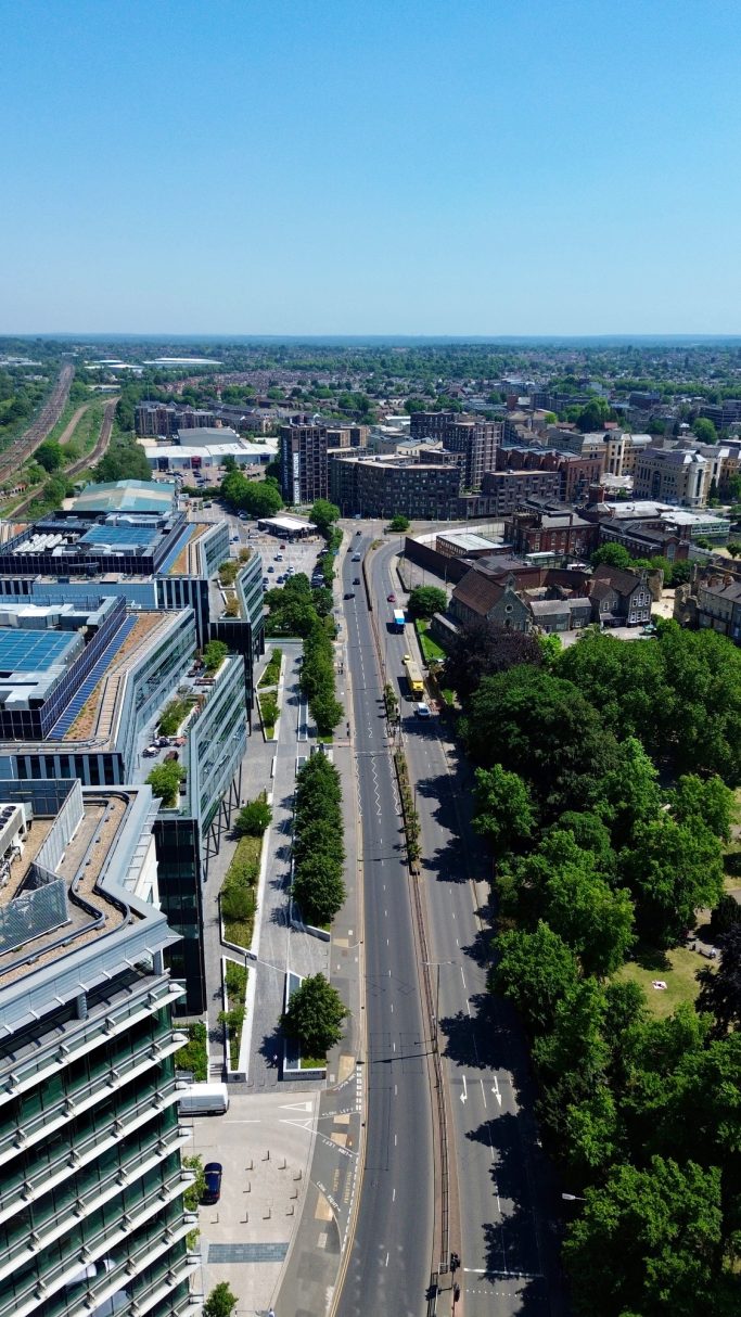 Aerial view of a road lined with trees and modern buildings under a clear blue sky.