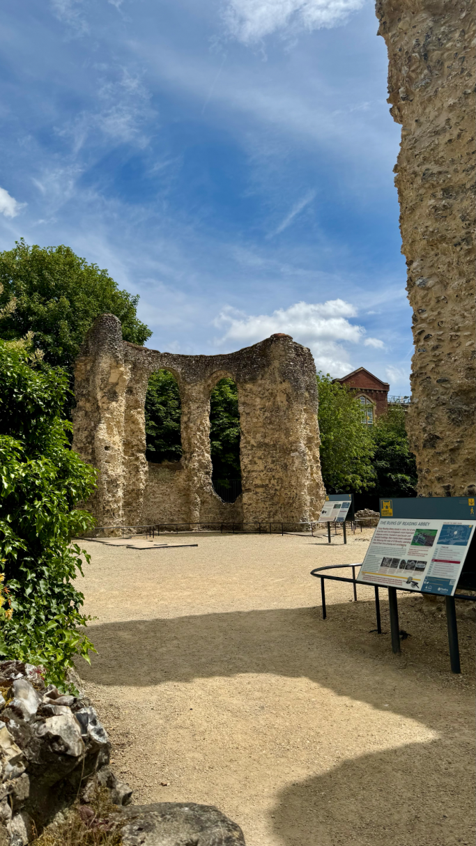 Ruins of stone walls with greenery and informational signs in a sandy area.